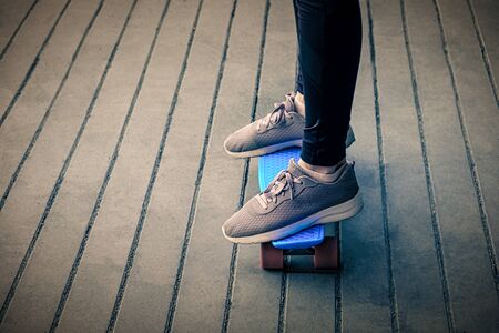 A girl whose feet are dressed in old sneakers stands on a bright blue skateboard on the pavement...