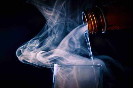 A brown bottle of alcohol hangs over a glass, and an evil spirit comes out of it and fills the glass with flow of liquer. A picture on a black background with a copypaste.の写真素材
