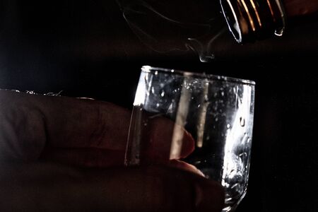 A man holds a glass in his hand over which the bottle's neck hangs and a trickle of steam flows out of it, tempting the evil spirit of alcohol.の写真素材