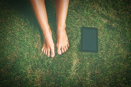 Tablet PC is lying on grass near the bare feet of female student. Shot from above. Faded look toned in vintage colours.の写真素材
