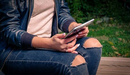 E-learning on-the-go. A girl in torn jeans sits on a park bench and prepares for exams using a...