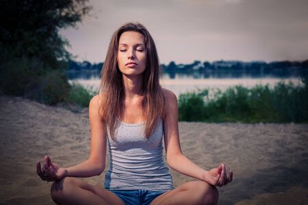 Blond haired lady is meditating in the park, seeking harmony and calmness inside. She is sitting cross legged in lotus-asana on the coastal sandの写真素材