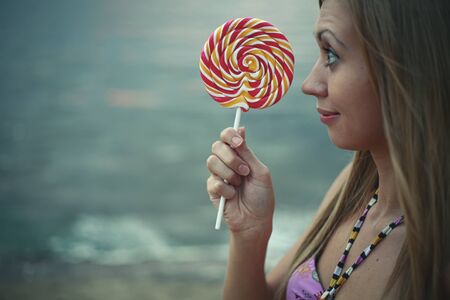 Girl is holding candy in front of her face in front of the sea wavesの写真素材