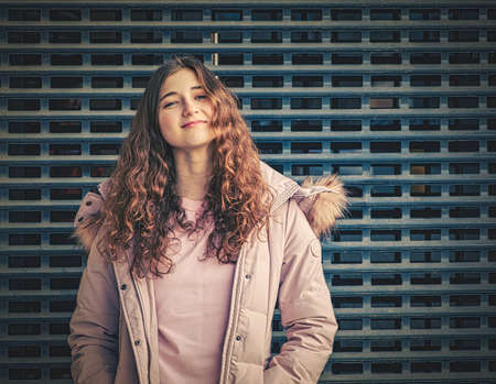 A redheaded girl with wavy hair poses in a jacket in front of a metal fence mesh. She puts her chin slightly up in the camera with confidence.の写真素材