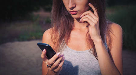 The young girl holds a mobile phone in her hands and looks at its screen on the beach, copyspace.の写真素材