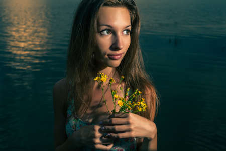 Young woman posing with bouquet of wildflowers on the sunset panorama of riverの写真素材