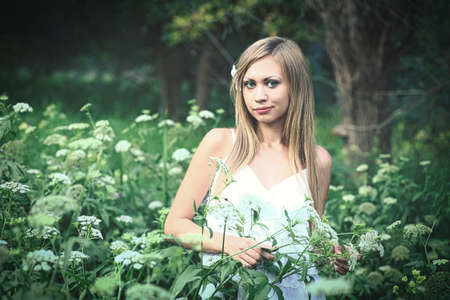 The nice blonde poses dressed in a white cloth amid the high grass and white wild flowersの写真素材