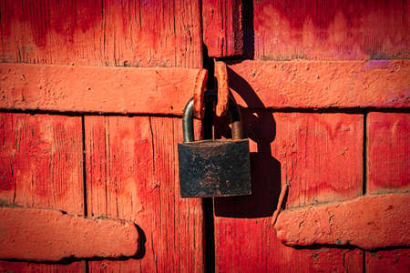 Closed lock on gates made of wooden planks and painted in red, retro looking image with shadows.の写真素材