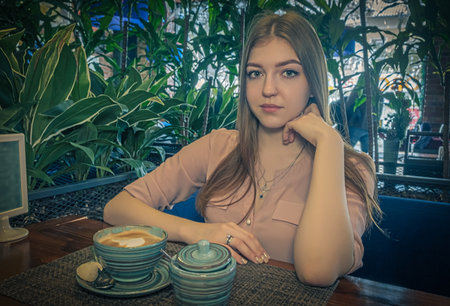Nice lady sitting in cafeteria with milk coffee with froth on top served in front on her with sugar bowl aside. She is looking at camera and touching her chin.の写真素材