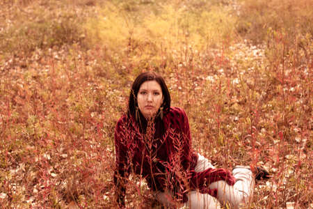 Sad dark-haired lady is sitting on the ground in tall autumnal grass, central composition.の写真素材