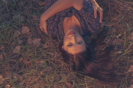 Top view of a gorgeous young lady closed eyes and wearing ornate pattern dress posing on the grass with autumn leaves. Beautiful female with closed eyes has dreamy expression lying outdoor in the park.の写真素材