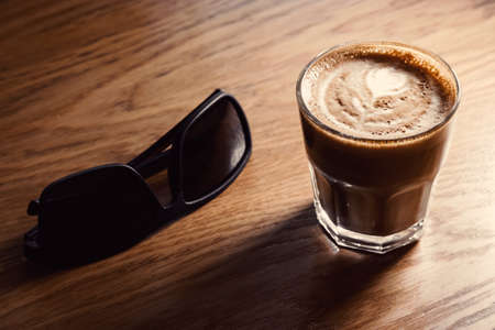 Sunglasses and flat-white coffee glass lying on wooden tableの写真素材