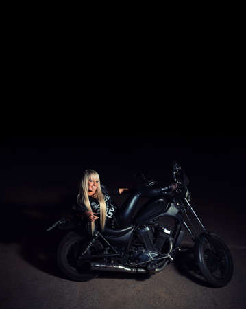 Young sexy fashionable blond haired lady lying on motorcycle bike in the night dark background, copyspace on topの写真素材