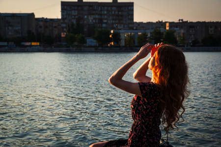 Portrait of a pensive young beautiful lady sitting on a river embankment parapet with city scape on other side in the background, shot with copyspace. Portrait of young redhead woman with long wavy hair relaxing in riverside.の写真素材