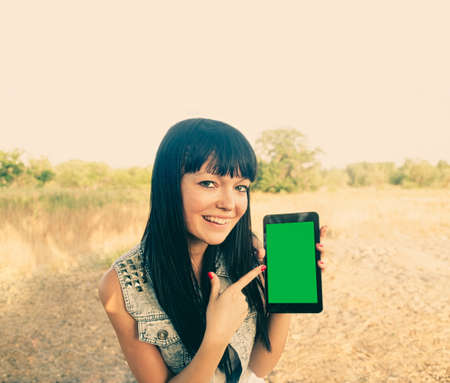 Brunette girl holds a tablet touch pad computer gadget with green screen outside in the summer and point at screenの写真素材