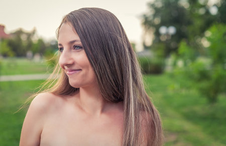 Photo of smiling millennial blond lady with bare shoulders posing outside in park with happy expression on her face looking aside camera.の写真素材