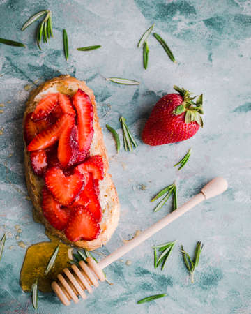 Sliced strawberries on toast surrounded by scattered rosemary, a whole strawberry, and a dripping...