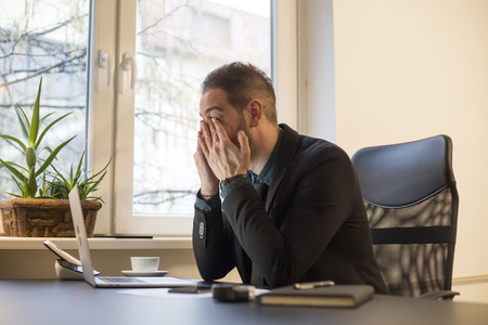 businessman working on laptop in office eyes hurt by window black table with notebook papers phoneの写真素材