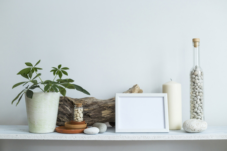 Modern room decoration with picture frame mockup. Shelf against white wall with decorative candle, glass, wood and rocks. Home plant in pot.の写真素材