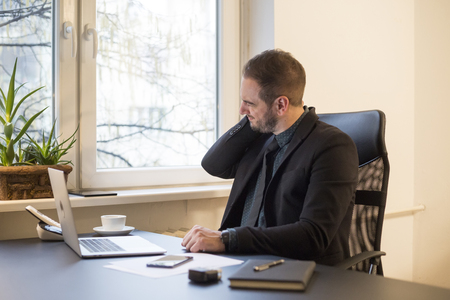 businessman working on laptop in office neck hurt by window black table with notebook papers phoneの写真素材