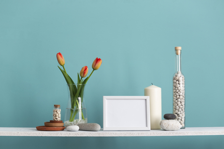 Modern room decoration with picture frame mockup. Shelf against turquoise wall with decorative candle, glass and rocks. Tulips in vase.の写真素材