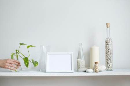 Modern room decoration with picture frame mockup. Shelf against white wall with decorative candle, glass and rocks. Hand putting down Plant cuttings in glass.の写真素材