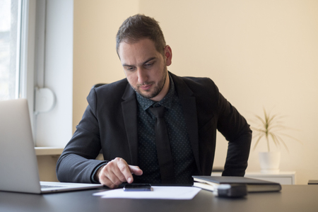 businessman in office sending messages with mobile phone smiling by window black table with notebook papers phone working laptopの写真素材