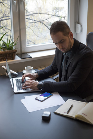 businessman working on laptop in office taking notes by window black table with notebook papers phoneの写真素材