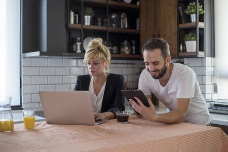Couple in kitchen in the morning. They are working on tablet and laptop. Small business start up...