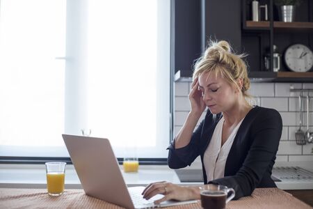 Young woman having headache while working on laptop. She is sitting at her kitchen table at home.