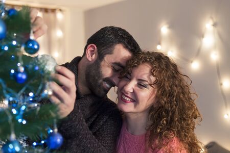Happy couple putting decoration on Christmas tree in living room. Holidays preparation.の写真素材