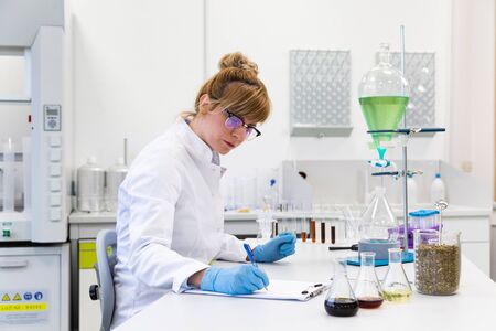 Female scientist wearing glasses and latex gloves writes measurement results with blue pen. Turquoise liquid in pear shaped separatory funnel is in background. Hemp seeds and cbd oils are on table.の写真素材