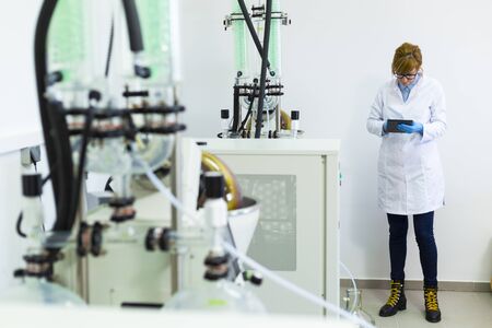 Scientist typing on black tablet in laboratory during CBDa oil extraction. She wears rubber gloves and stands next to rotational vaporizer with green condenser called rotavapor.の写真素材