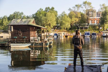Man with long hair is fishing at river corner from raft. Boats and floating houses are around him with trees in background. He is using fishing rod with spinning reel, green line and lure.の写真素材