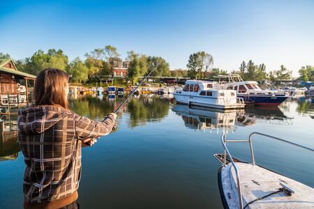 Man with long hair is fishing at river corner from raft. Boats and floating houses are around him with trees in background. He is using fishing rod with spinning reel, green line and lure.の写真素材