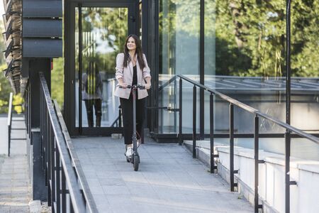 A young beautiful girl is riding her electric scooter. She is smiling, business-dressed, with modern...