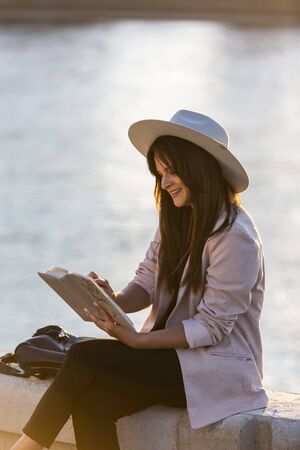 A young beautiful girl with a hat is sitting by the river with the book in her hands. The river and a bridge predominate in the background.の写真素材