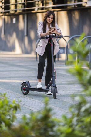 A young beautiful girl standing next to electric scooter while typing on her phone. She is smiling,...