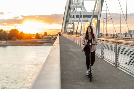 A young beautiful girl is riding her electric scooter over a bridge. She is business-trained. The background consists of the parts of a modern bridge.の写真素材