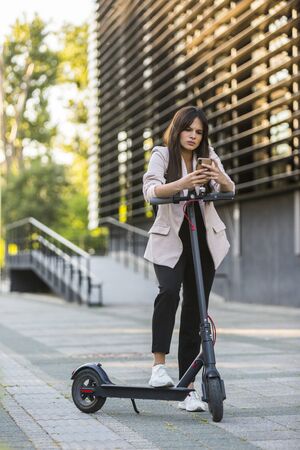 A young beautiful girl standing next to electric scooter while typing on her phone. She is smiling, business-dressed, with modern architecture in the background.の写真素材