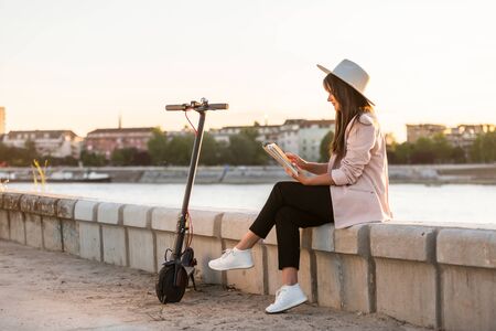 A young beautiful girl with a hat is sitting by the river with the book in her hands. An electric scooter is parked next to her, while the river and a bridge predominate in the background.の写真素材