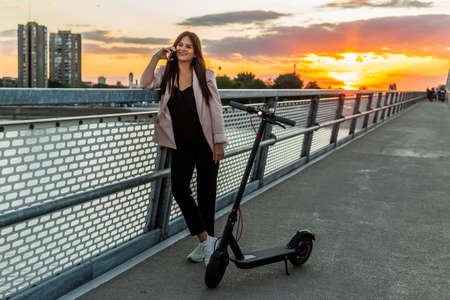 A young smiling woman is standing by the bridge fence while talking on her phone. An electric scooter is parked next to her, while the buildings, sunset and a bridge fence predominate in the backgroundの写真素材