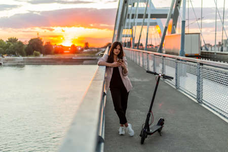 A young smiling woman is leaning on the bridge fence and typing on her phone. An electric scooter is parked next to her. River, sunset and the parts of a modern bridge predominate in the background.の写真素材