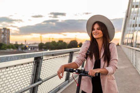 A young beautiful woman is standing next to her electric scooter with one foot on a standing platform. She is wearing a hat and she is business-trained. The background consists of the parts of a bridgeの写真素材