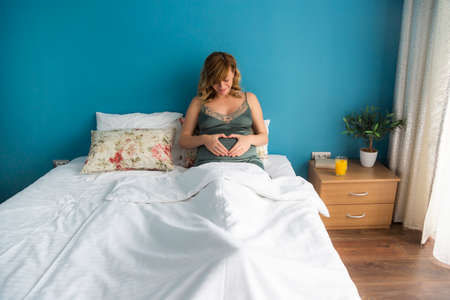 A smiling pregnant woman in a semi-sitting position in bed with her hands around her stomach. She's looking down at her stomach.の写真素材