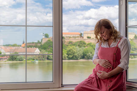 Happy pregnant caucasian woman sitting by the window while part of the old town and the river are in the background. Her gaze is directed downward as she holds her arms around her stomach.の写真素材