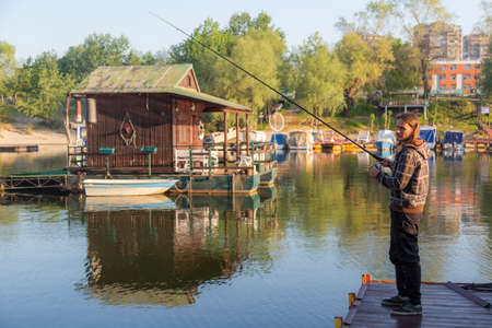 Man with long hair is fishing at river corner from raft. Boats and floating houses are around him with trees in background. He is using fishing rod with spinning reel, green line and lure.の写真素材