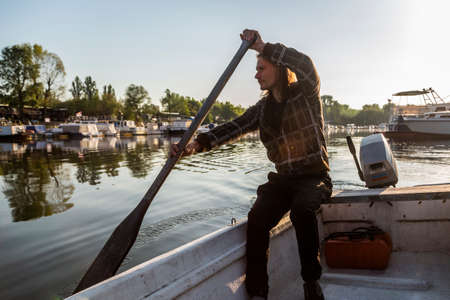 White mid age man rowing on his old white boat. He is in river marina at sunrise. For oarage this fisherman is using old wooden oar. Boats and floating houses are around him with trees in background.の写真素材