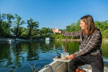 Fisherman throwing chum in lake. He is in white boat with trees around him. Guy has long brown hair. Construction yard is in background. Corn and worms splashing on water surface causing drops.の写真素材