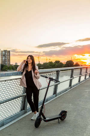 A young smiling girl is standing by the bridge fence while talking on her phone. An electric scooter is parked next to her, while the buildings, sunset and a bridge fence predominate in the backgroundの写真素材